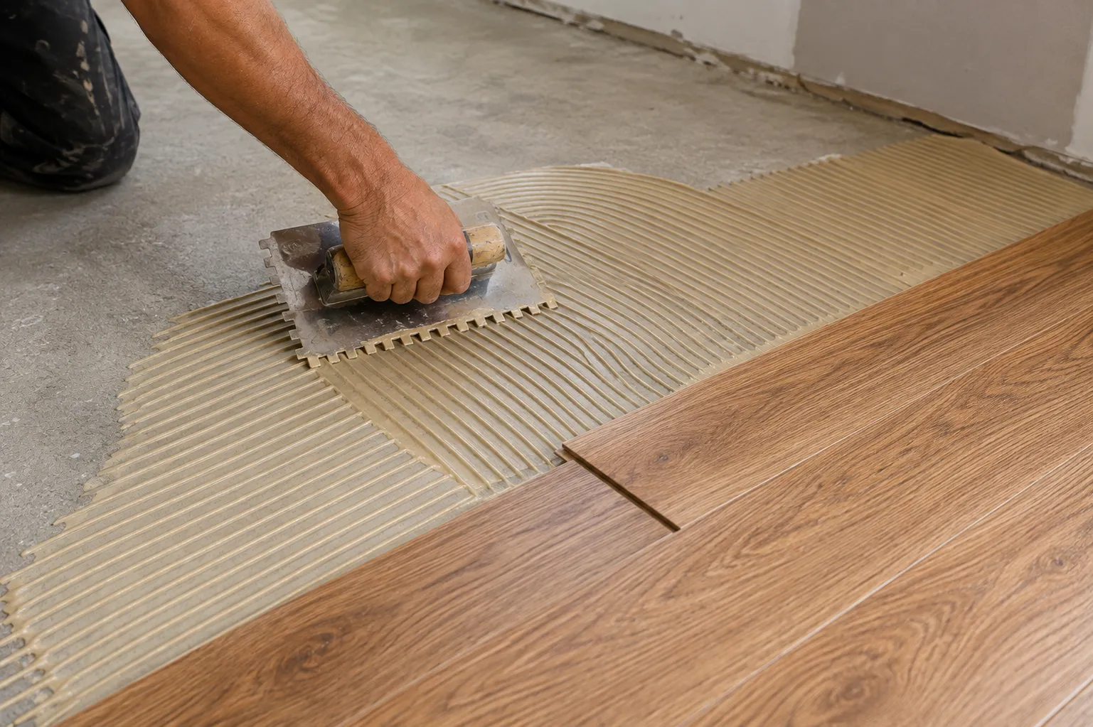 Notched trowel spreading flooring adhesive over a concrete slab before laying engineered planks